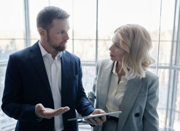 Two business professionals in formal attire engaged in a focused workplace discussion, holding a tablet.