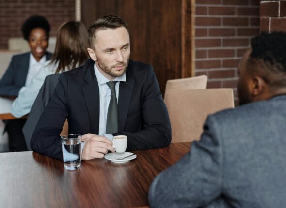 A business professional in a suit holds an espresso while listening intently to a colleague, with two other professionals conversing in the background.