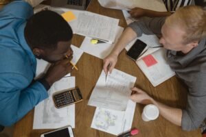 Two professionals working together at a table, reviewing documents, charts, and a calculator, illustrating teamwork and effective communication.